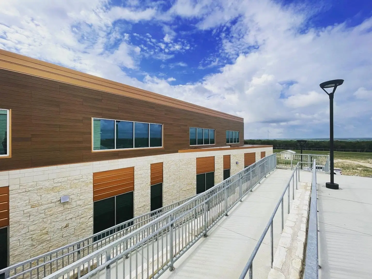 A two-story Dripping Springs ISD campus building in cream limestone and wood-grain panel, a ribbon of windows above and an accessible ramp running along the front, with open Hill Country beyond