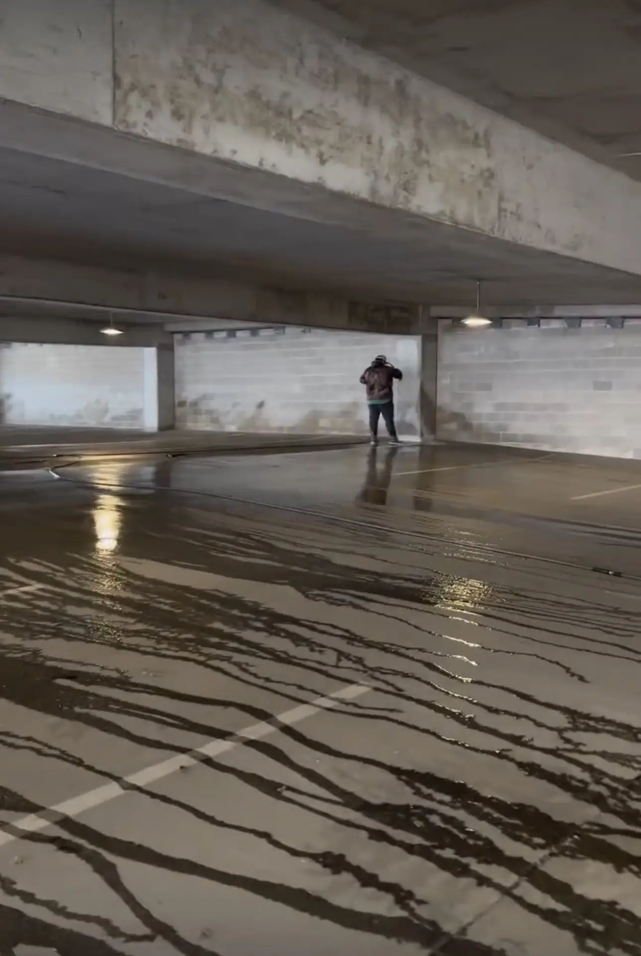 An operator working a parking deck level with a wand, dark runoff streaking in long lines across the wet concrete toward the camera