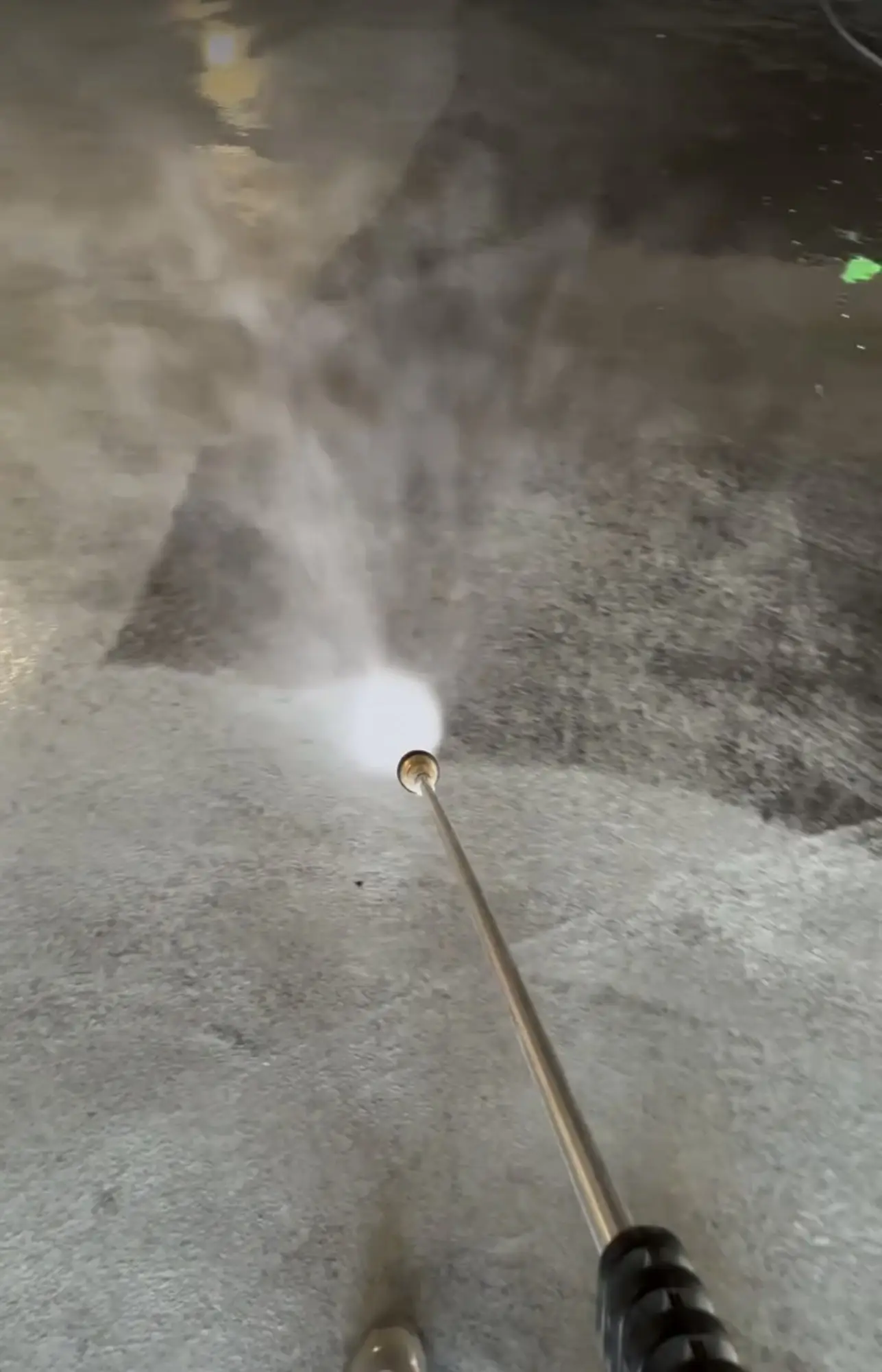 A close view of a pressure washing wand lifting dirt from a concrete deck, a pale cleaned fan opening across the darker surrounding surface