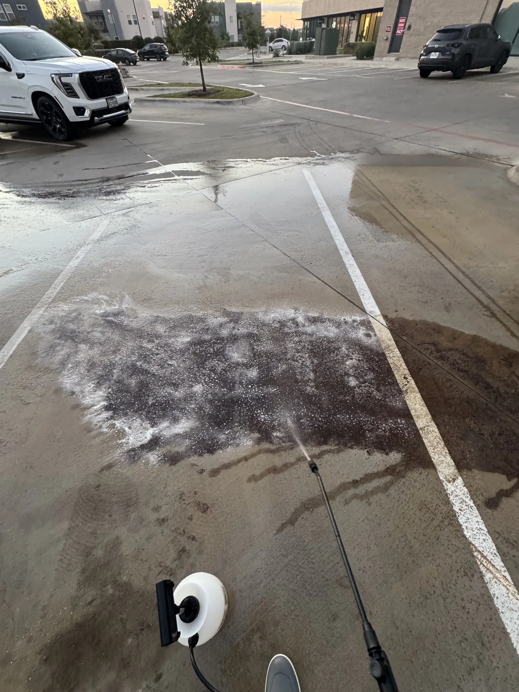Degreaser being applied with a wand across the oil-stained parking spaces as the sun rises behind the shopping center buildings