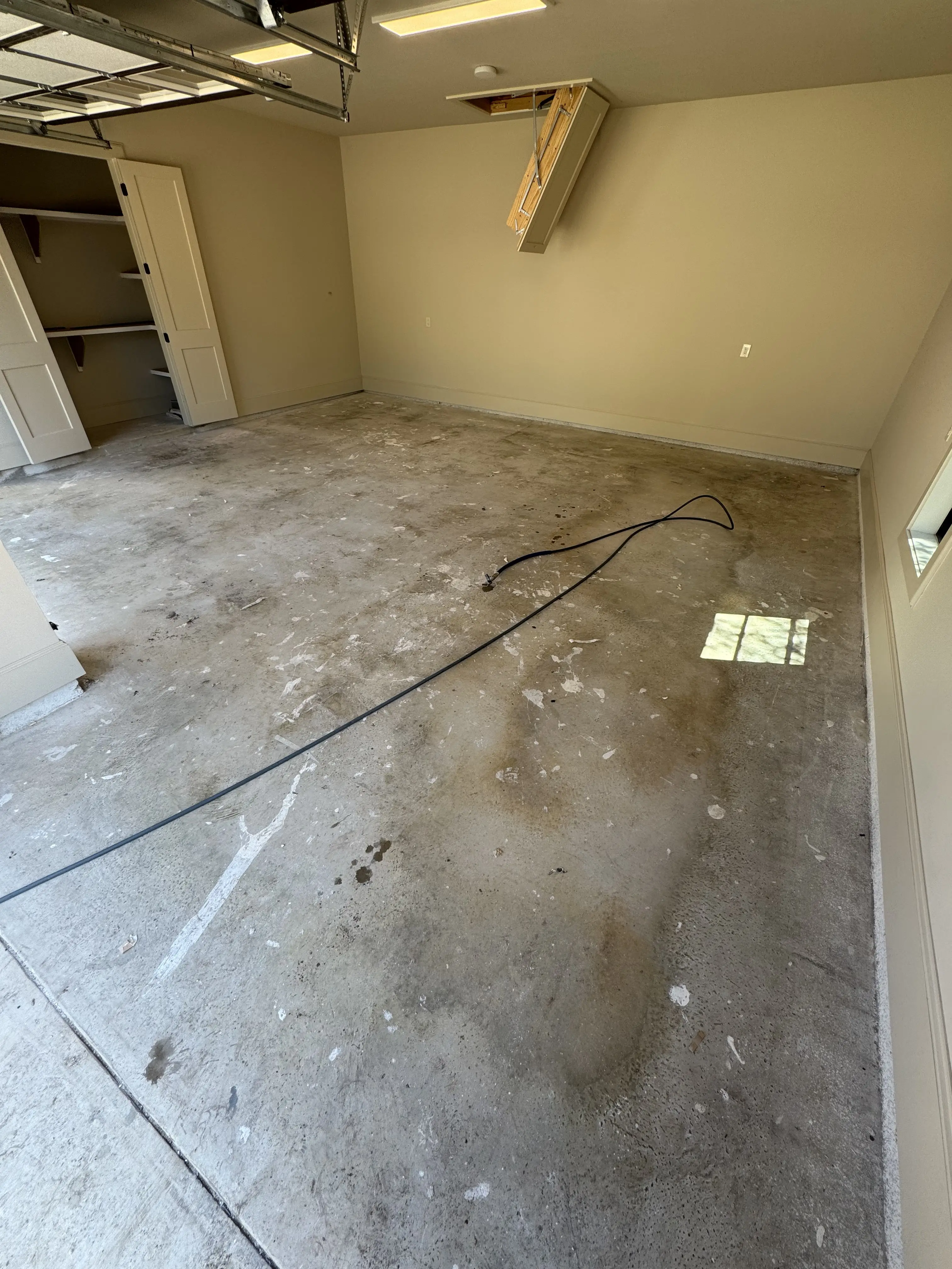 A new garage floor before cleaning, white drywall mud splatter and dark traffic staining across the slab, the attic ladder down and the garage door open at the left