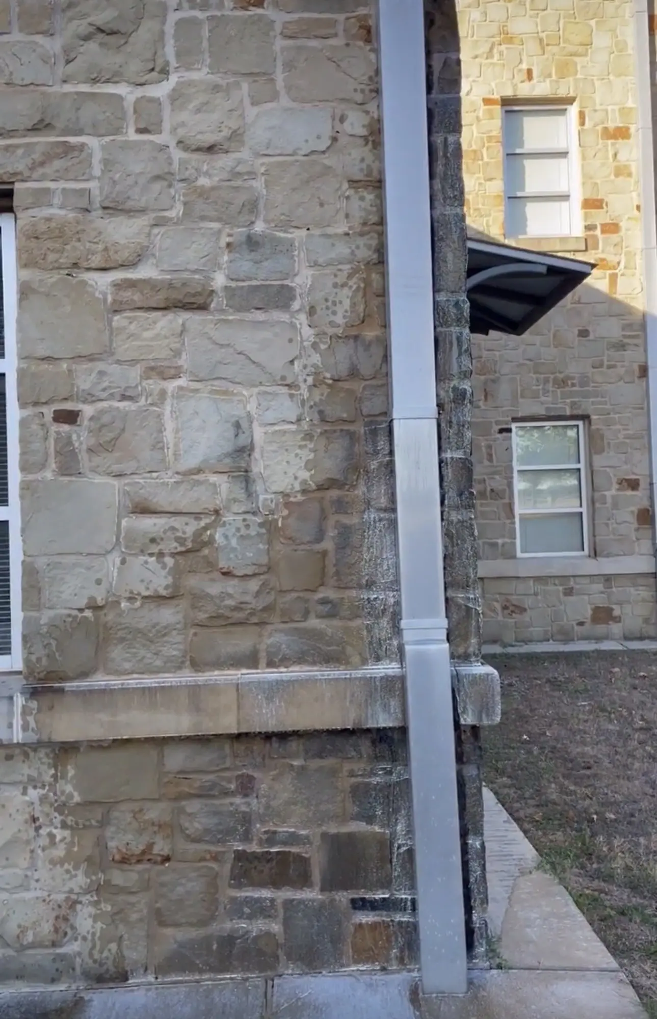 A limestone campus wall with dark organic staining running down the stone beside a downspout, the cleaner pale stone of the next elevation visible behind