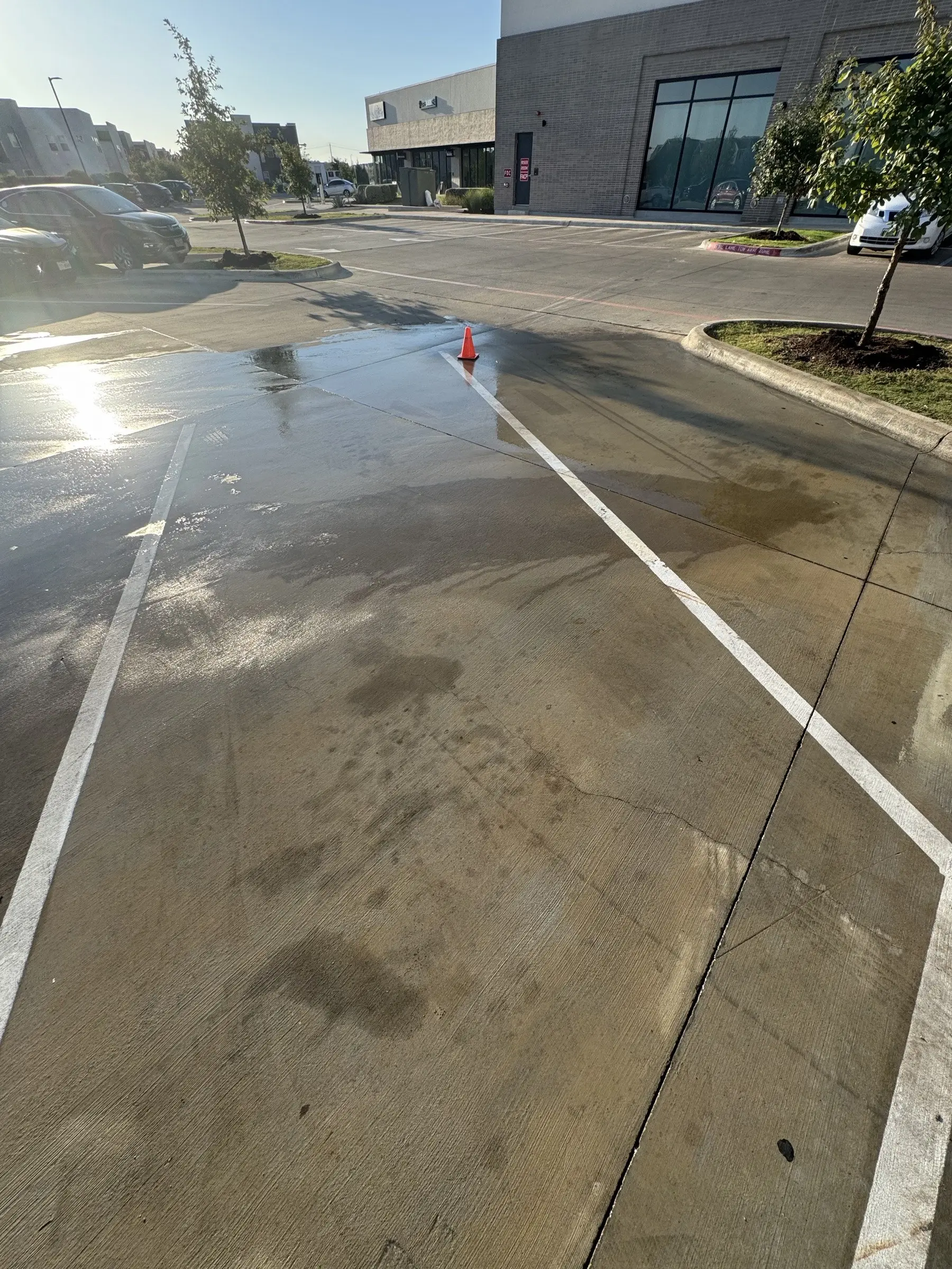 The cleaned parking spaces wet and back in service at sunrise, a safety cone at the edge and the shopping center storefronts behind