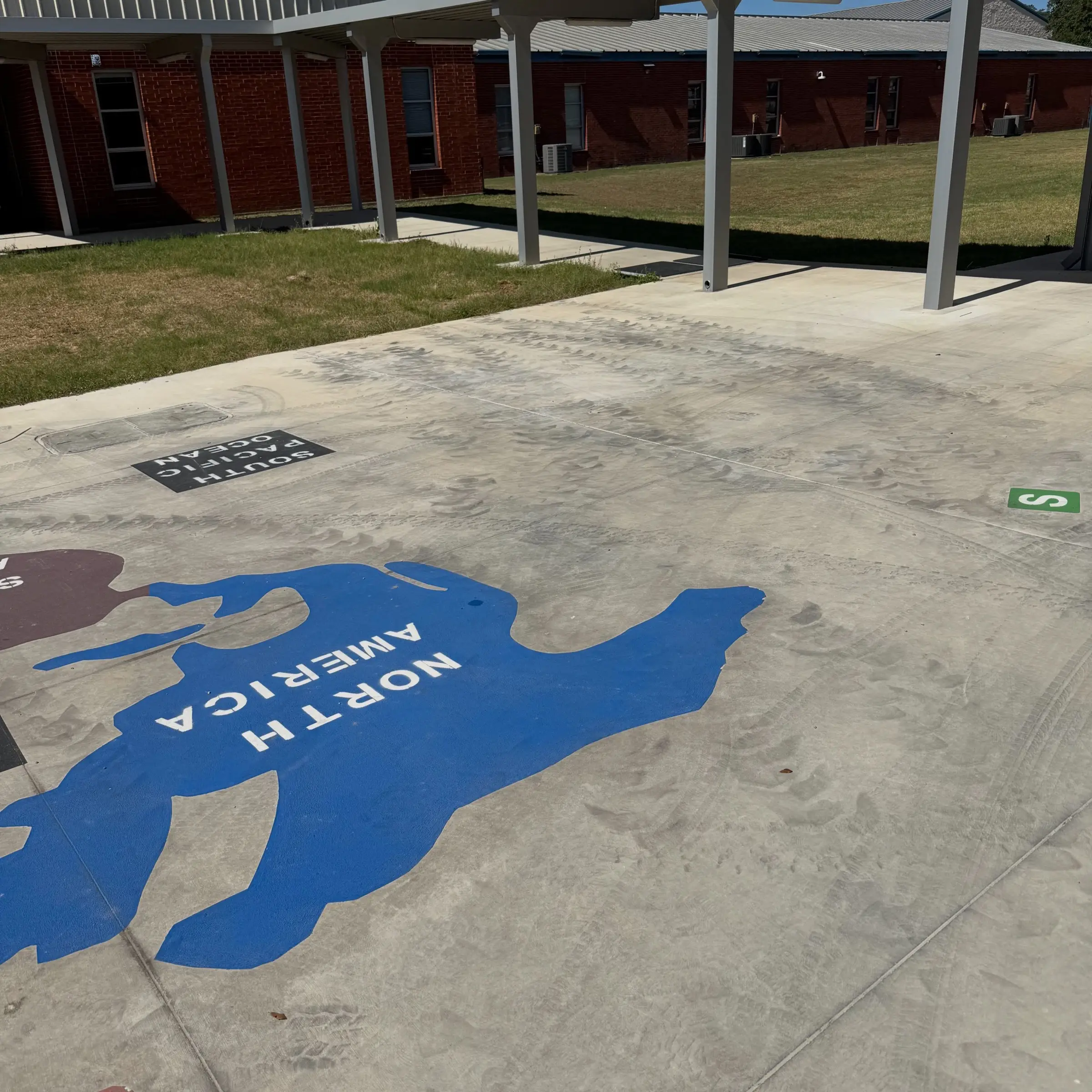 Construction tire tread marks arcing across the painted world map play surface at a Blanco ISD campus, with the red brick school and covered walkway behind